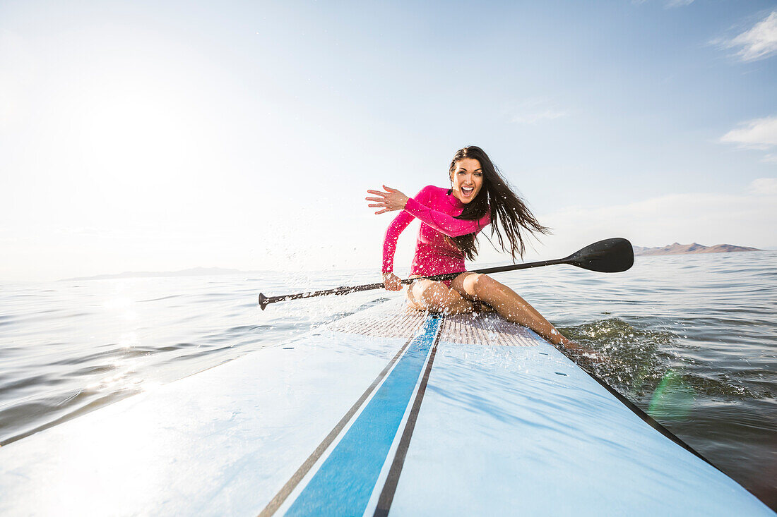 Lächelnde Frau in rosa Badeanzug auf Paddleboard sitzend und Wasser spritzend