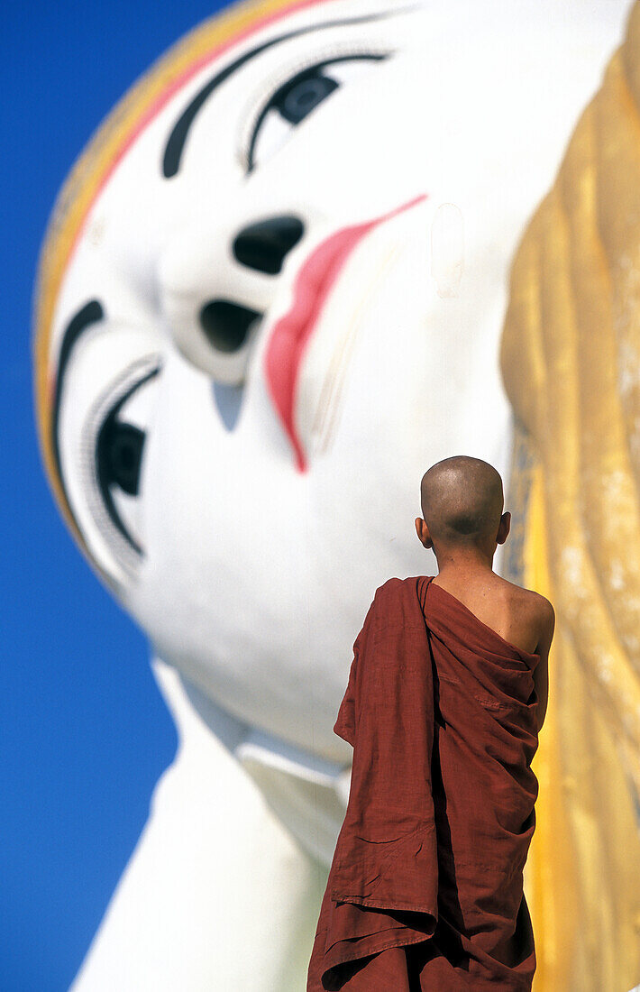 Myanmar, Monyma, Mandalay Division, Novice monk praying under giant statue of reclining Buddha in Lay Kyune Sakkyar temple