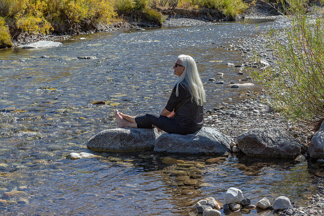 USA, Idaho, Sun Valley, Ältere blonde Frau sitzt auf einem Felsen am Bach 