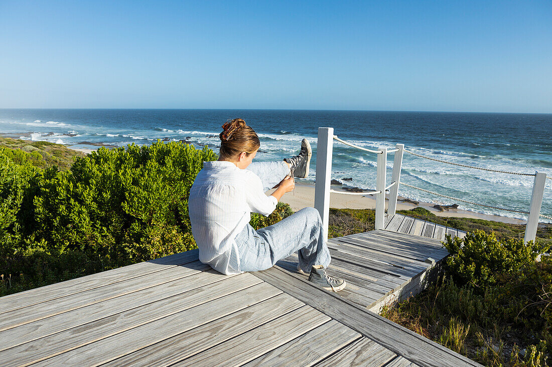 South Africa, Western Cape, Girl (16-17) sitting on boardwalk and putting shoes on in Lekkerwater Nature Reserve