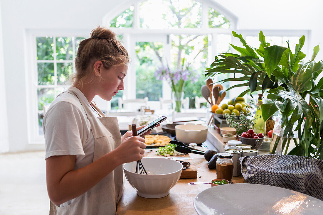 Girl (16-17) using phone while preparing meal in kitchen