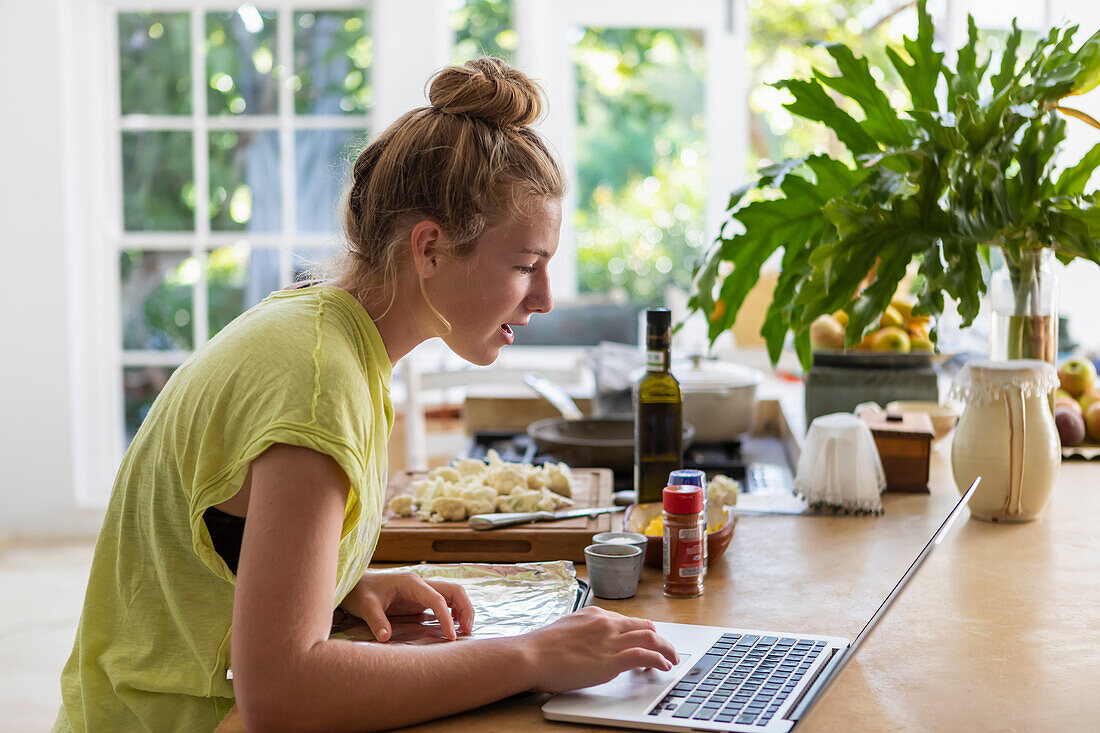 Mädchen (16-17) benutzt Laptop bei der Zubereitung einer Mahlzeit in der Küche