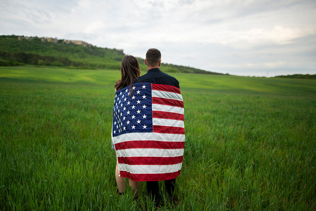 Rückansicht eines in die amerikanische Flagge gehüllten jungen Paares in einem Weizenfeld