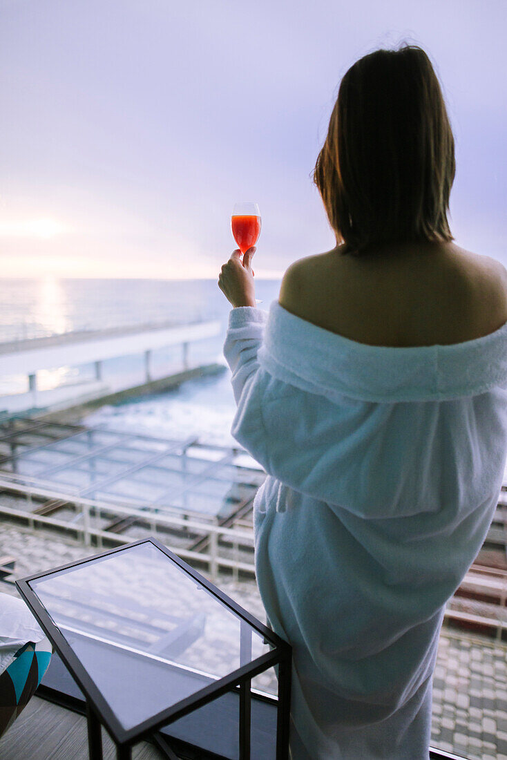 Rückansicht einer Frau mit Cocktail im Hotel und Blick auf den Sonnenaufgang über dem Meer