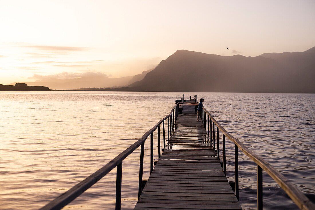 South Africa, Stanford, Boy (10-11) standing on pier at lagoon at sunset