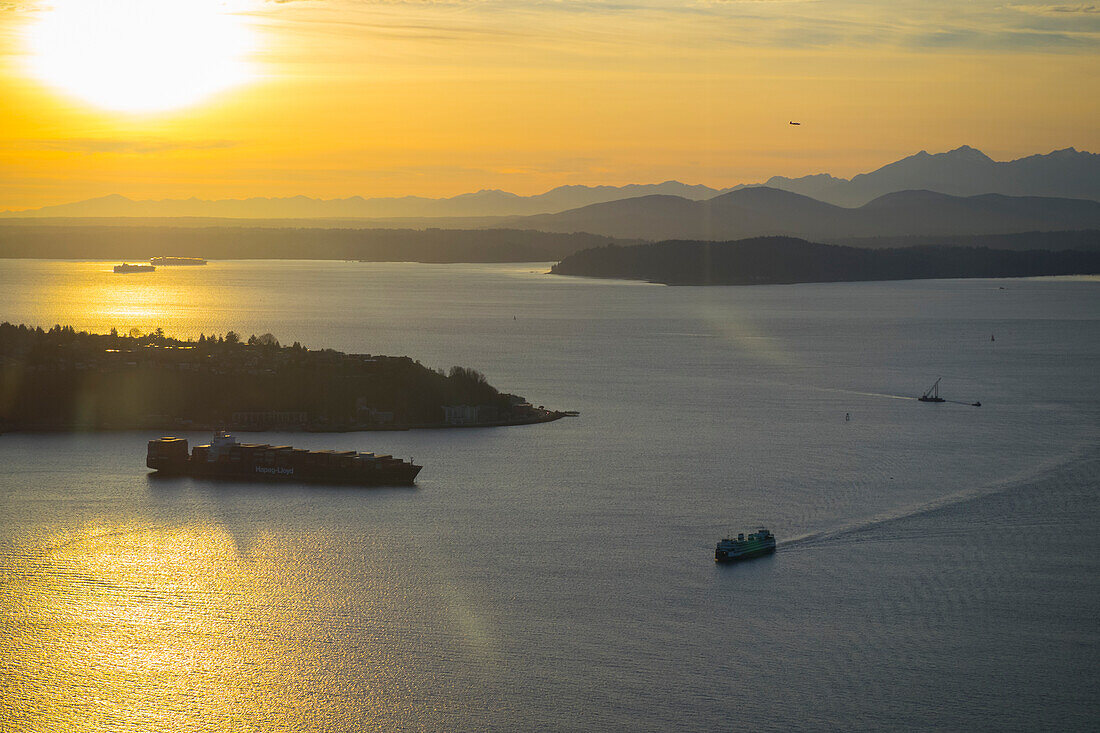USA, Bundesstaat Washington, Seattle. Eine Fähre des Staates Washington überquert den Puget Sound bei Seattle, Sonnenuntergang. Ein Containerschiff passiert West Seattle auf der linken Seite und die Olympic Mountains im Hintergrund.