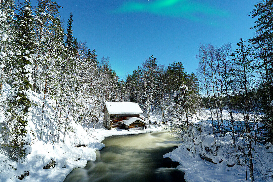 Aurora Borealis (Nordlicht) über einer Hütte mit Blick auf die Stromschnellen des zugefrorenen Flusses Myllikoski, Ruka Kuusamo, Lappland, Finnland, Europa