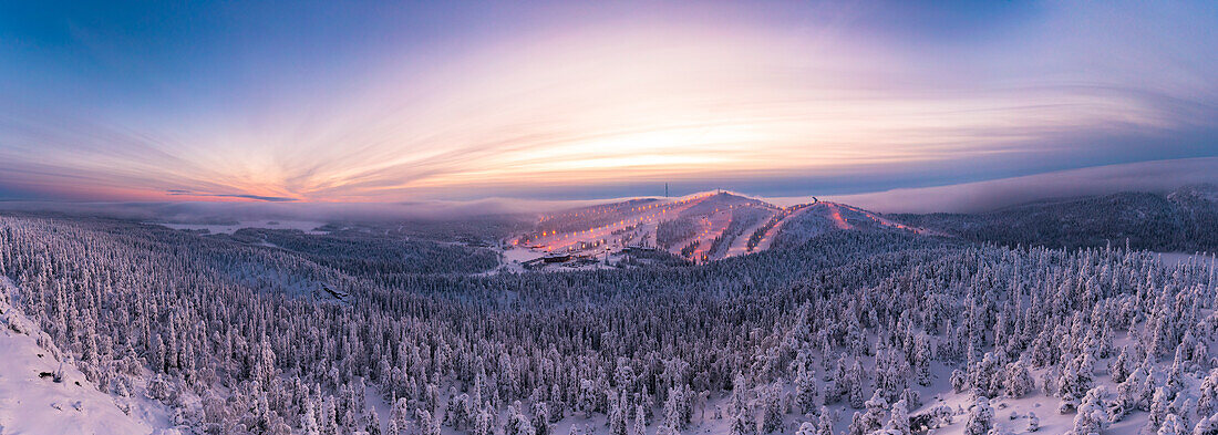 Panoramablick auf verschneite Wälder und Skipisten unter dem dramatischen Himmel bei Sonnenuntergang, Ruka, Kuusamo, Nordösterbotten, Lappland, Finnland, Europa