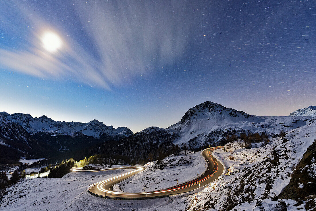Der Vollmond leuchtet über einer verschneiten Bergstraße, die von Autospuren beleuchtet wird, Berninapass, Val Poschiavo, Kanton Graubünden, Schweiz, Europa