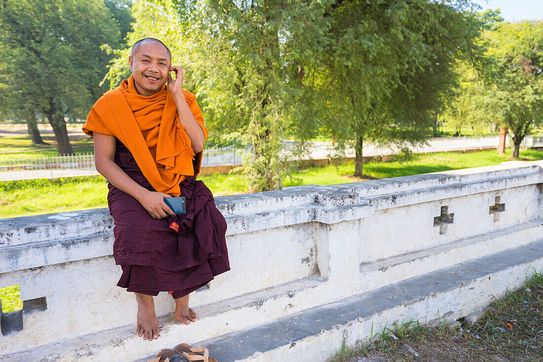 Mönch in safranfarbener Robe sitzt mit seinem Handy in der Hand an der Wand, Königspalast, Mandalay, Myanmar (Burma), Asien