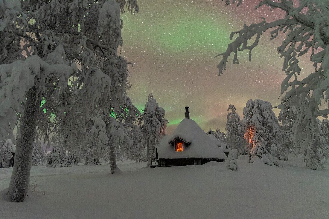 Typische beleuchtete Holzhütte unter den Nordlichtern (Aurora Borealis) im frostigen Wald mit schneebedeckten Bäumen, Pallas-Yllastunturi-Nationalpark, Muonio, Lappland, Finnland, Europa