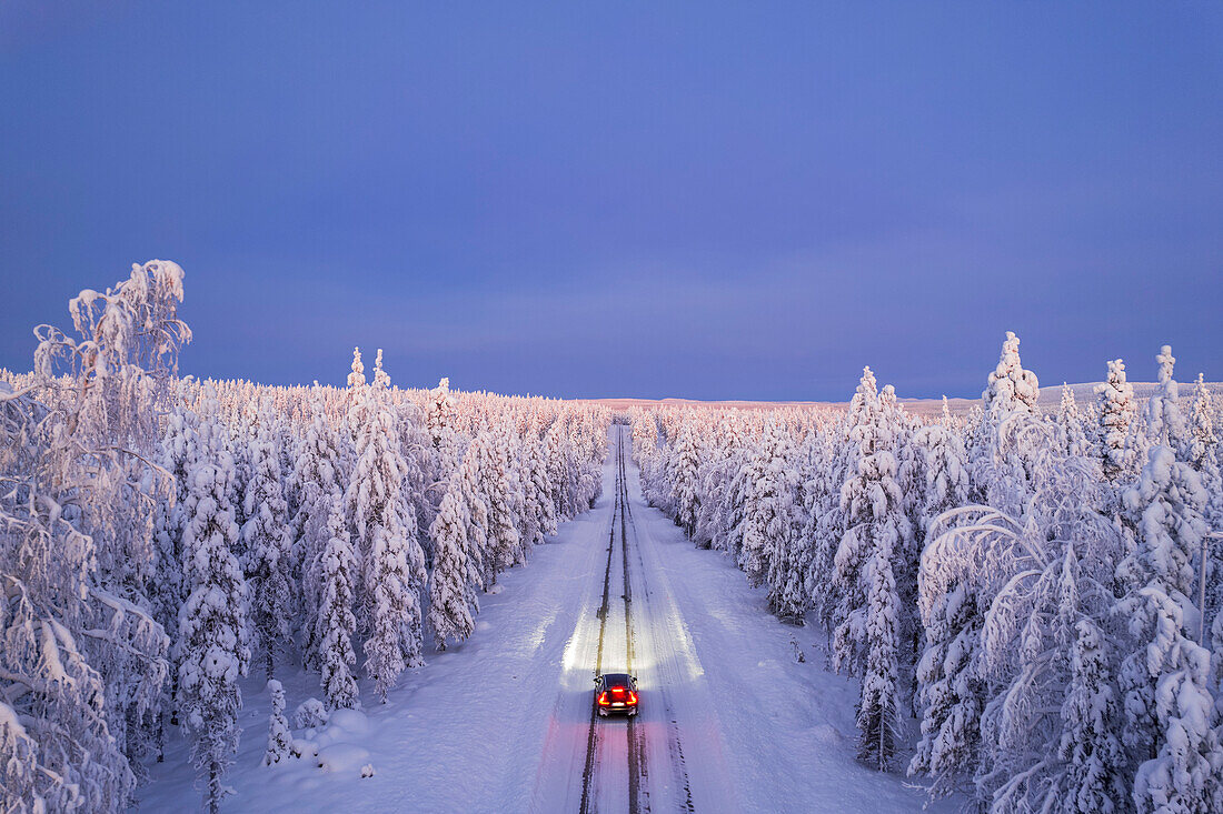 Blick von oben auf ein Auto, das auf leerer, eisiger und glatter Straße mit beleuchteten Scheinwerfern fährt, Akaslompolo, Kolari, Pallas-Yllastunturi-Nationalpark, Region Lappland, Finnland, Europa