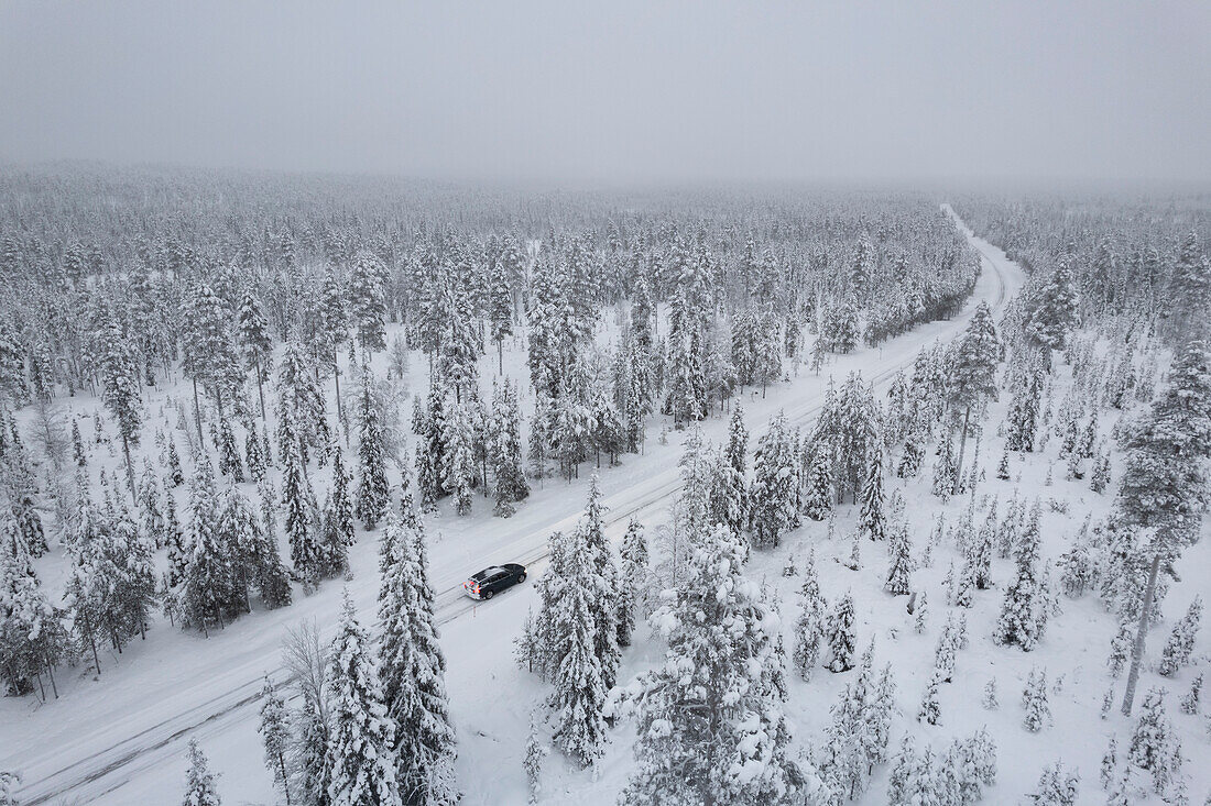 Blick von oben auf ein Auto, das auf einer vereisten Straße inmitten des gefrorenen, schneebedeckten Waldes fährt, Akaslompolo, Kolari, Pallas-Yllastunturi-Nationalpark, Region Lappland, Finnland, Europa