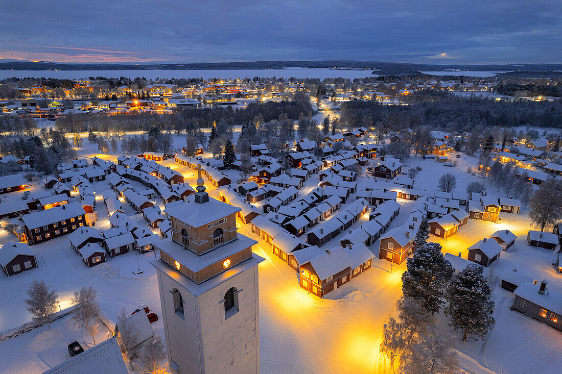 Luftaufnahme von beleuchteten Häusern und dem mit Schnee bedeckten Glockenturm im alten Dorf Gammelstad Church Town in der Abenddämmerung, UNESCO-Weltkulturerbe, Lulea, Norrbotten, Norrland, Schweden, Skandinavien, Europa