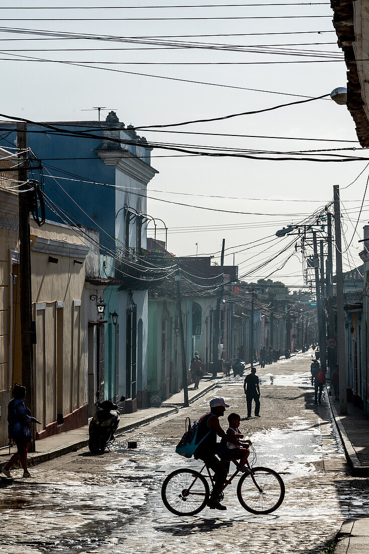 Typische Seitenstraße unter einem Gewirr von Telefonkabeln, Silhouette einer Familie auf dem Fahrrad, Trinidad, Kuba, Westindien, Karibik, Mittelamerika