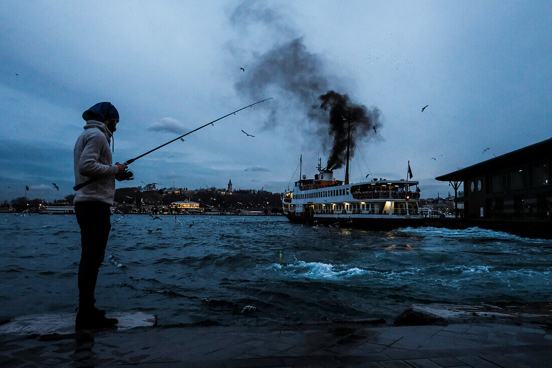Man fishing in the Golden Horn in Galata neighbourhood while a ferry boat is sailing