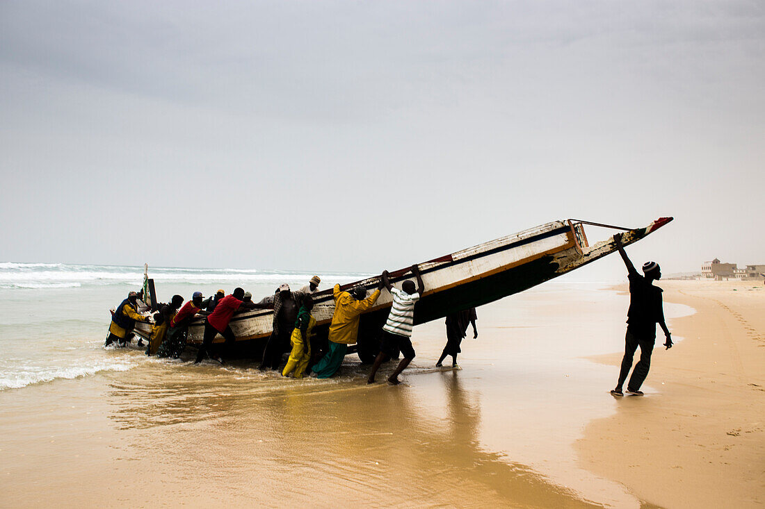 Männer entladen ein Fischkanu auf dem nachmittäglichen Fischmarkt am Strand von Mboro Plage, wenn die Kanus beladen ankommen