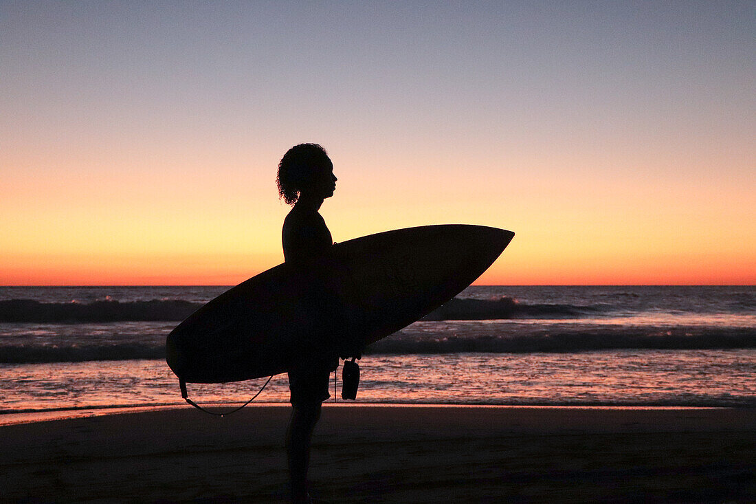 Silhouette of Oniel Lopez, young male surfer holding his surfboard at sunset on Jiquilillo beach, Nicaragua