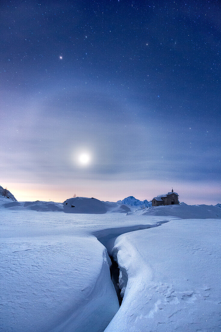 Moon halo in the night sky above a traditional mountain church during a winter full moon night. Valmalenco, Valtellina, Lombardy, Italy, Europe