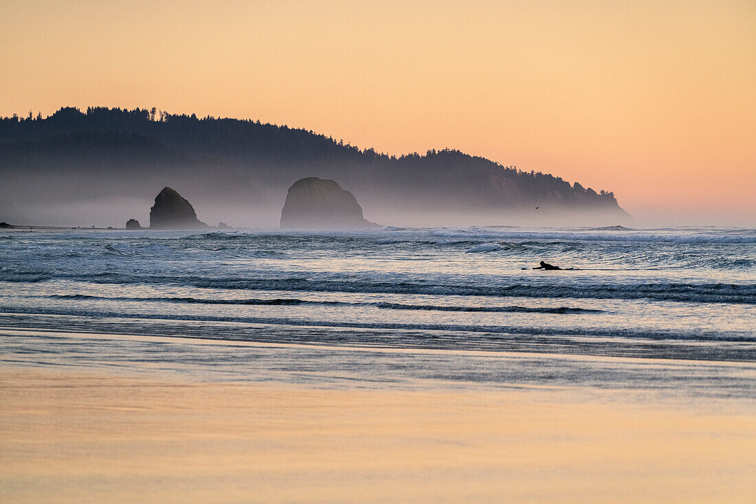Eine Person beim Surfen im Pazifischen Ozean bei Sonnenuntergang. Cannon Beach, Clatsop County, Oregon, USA.