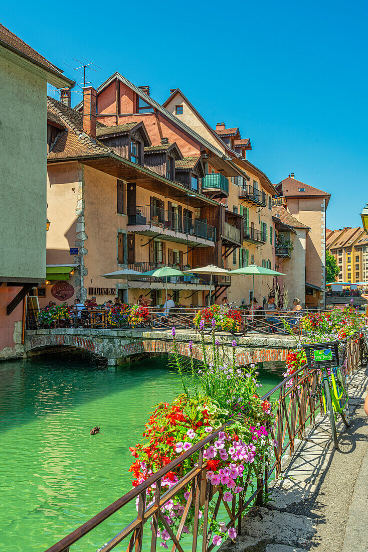 Tourists sit at tables in a cafe on one of the bridges that cross the Thiou River in the old town of Annency. Annecy, Savoie department, France