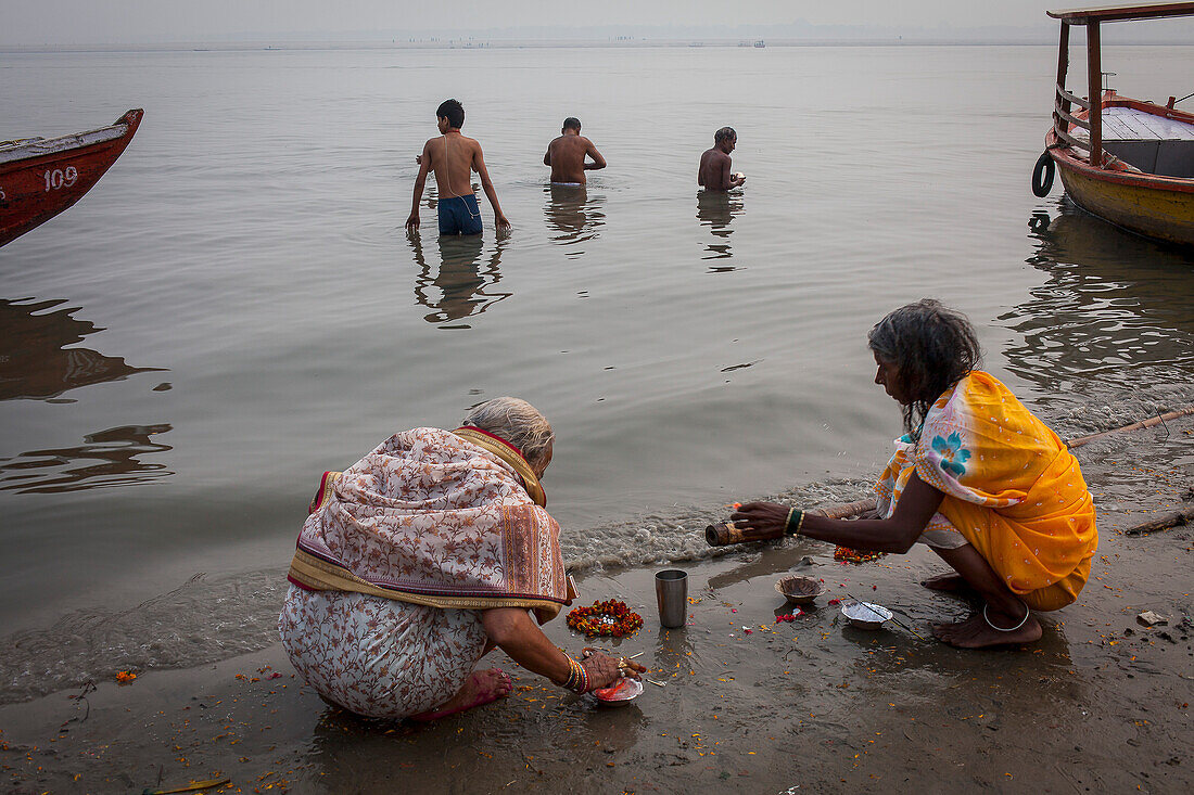 Frauen, die beten und Opfergaben darbringen, im Hintergrund Männer beim Baden, Ghats des Ganges, Varanasi, Uttar Pradesh, Indien.