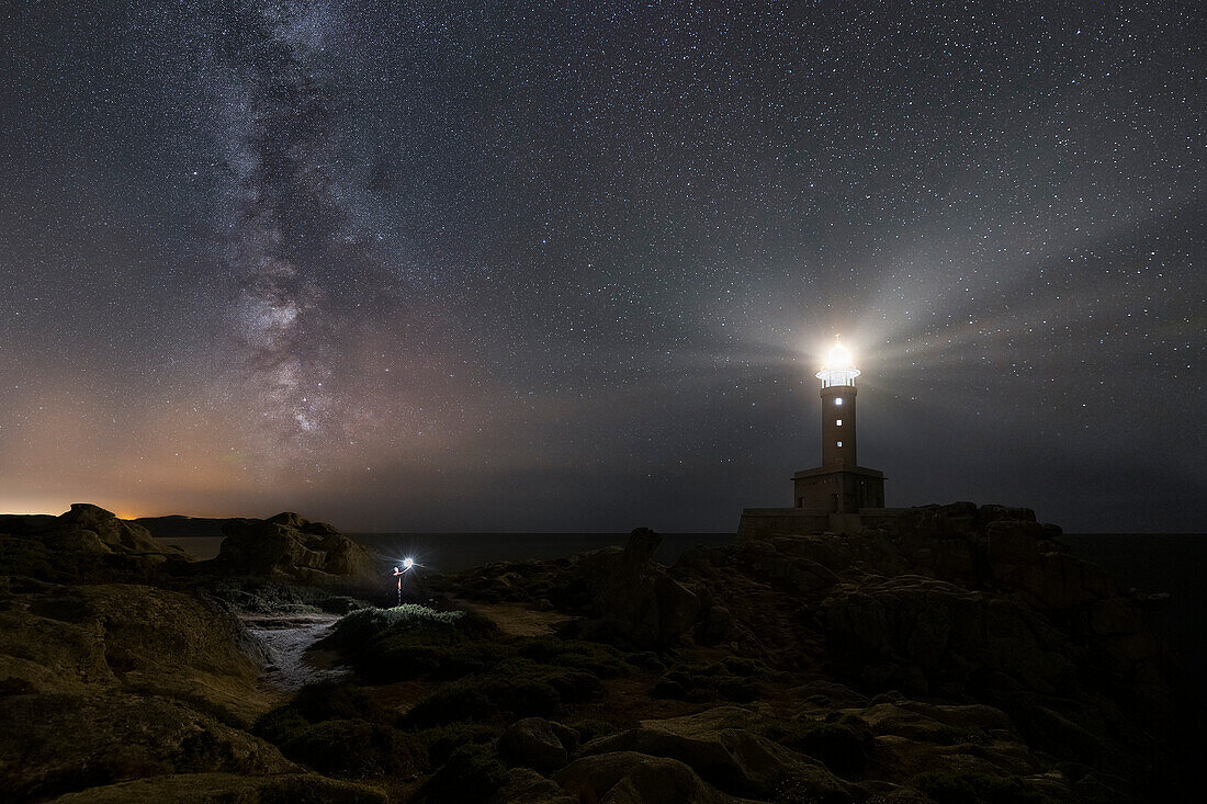 Ein Mann mit einer Taschenlampe beobachtet einen beleuchteten Leuchtturm von Punta Nariga bei Nacht mit Milchstraße, Costa da Morte, Galicien, Spanien, Iberische Halbinsel, Westeuropa