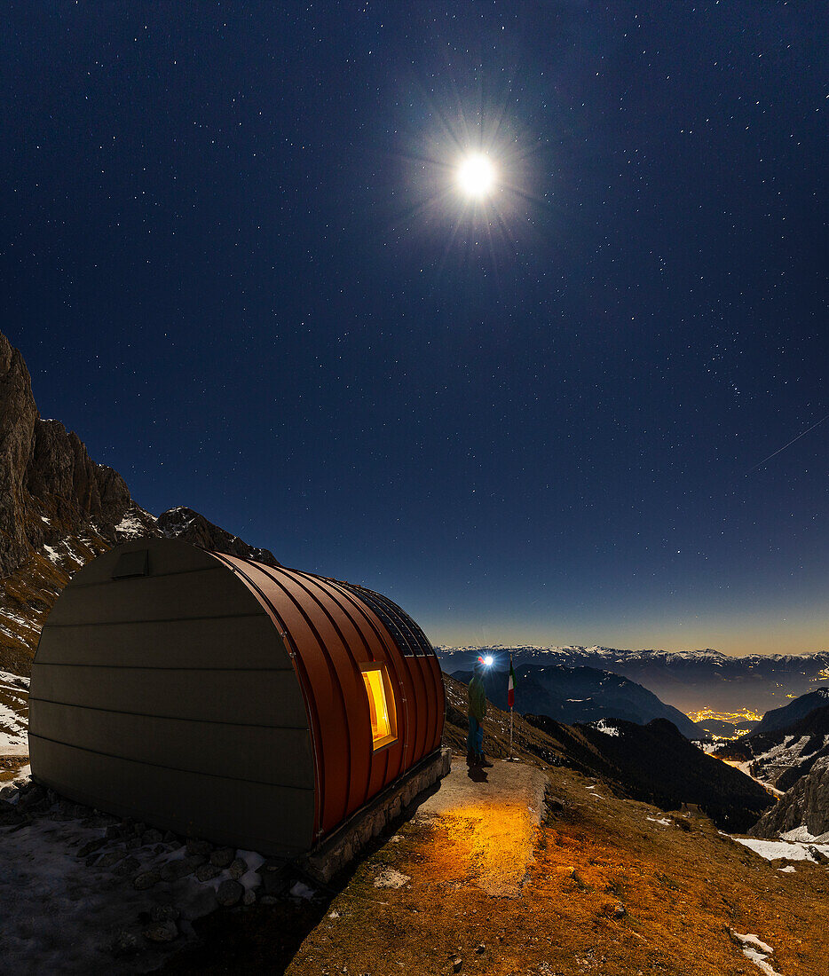 Ein Wanderer kommt aus dem beleuchteten Biwak Città di Clusone während der Nacht, Castione della Pesolana, Prealpi Orobie, Bergamo, Lombardei, Italien, Südeuropa