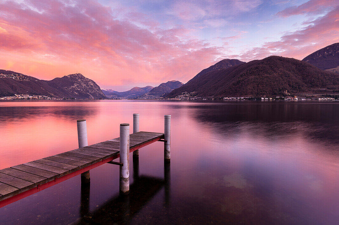 Explosion der Farben bei Sonnenaufgang vor Lugano und Campione d'Italia von einem Pier am Ceresio-See, Riva San Vitale, Kanton Tessin, Schweiz.