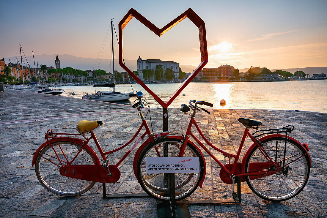 Toscolano maderno gulf, with position for touristic photos, red bike with heart, church of St. Andrea, boats, with sun at sunrise, Lake of Garda, Toscolano Maderno, Brescia province, Lombardy, Italy, north Italy, Europe, south Europe