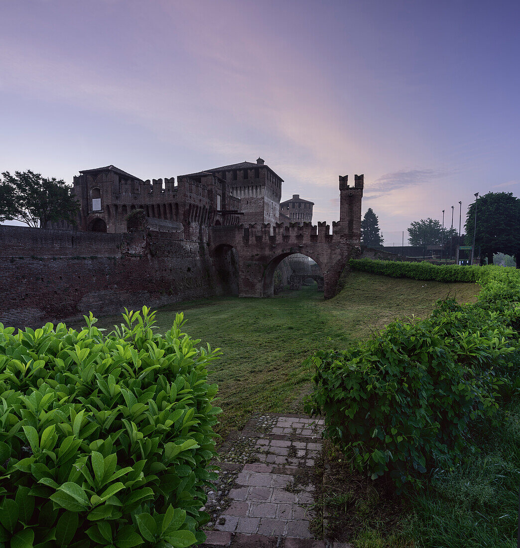 Schloss von Soncino, morgendlicher Seiteneingang mit Brücke, Vegetation und rotem Himmel, senkrecht Soncino, Provinz Cremona, Lombardei, Italien, Europa