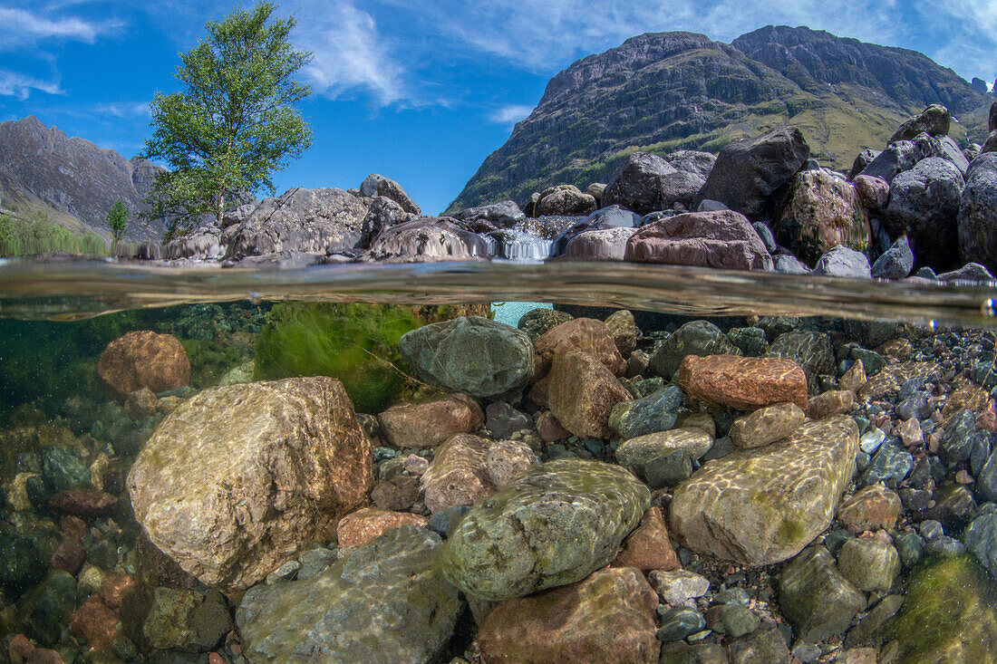 Rocks and pebbles under the River Coe on a sunny Scottish day. The three sisters mountains, blue skies and a tree are all visible above the water line, Glencoe, Scotland.