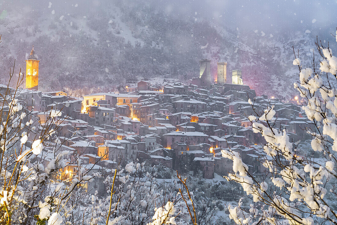 Abenddämmerung im beleuchteten mittelalterlichen Dorf Pacentro von unten gesehen unter starkem Schneefall mit dem Schloss und dem Glockenturm im Hintergrund und dem schneebedeckten Haus, Gemeinde Pacentro, Nationalpark Maiella, Provinz L'aquila, Abruzzen, Italien