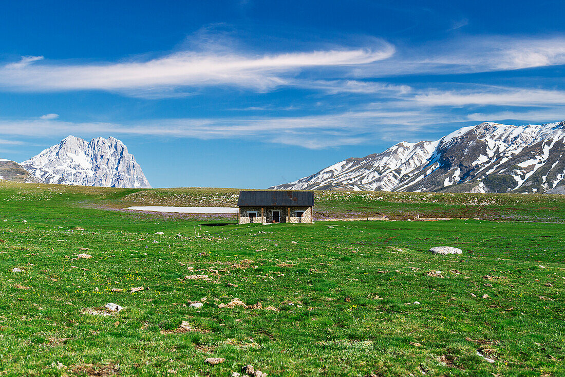 Einsames Haus und grüne Wiesen auf der Hochebene von Campo Imperatore mit dem Gipfel des Gran Sasso im Hintergrund im Frühling, Nationalpark Gran Sasso und Monti della Laga, Provinz L'Aquila, Abruzzen, Italien