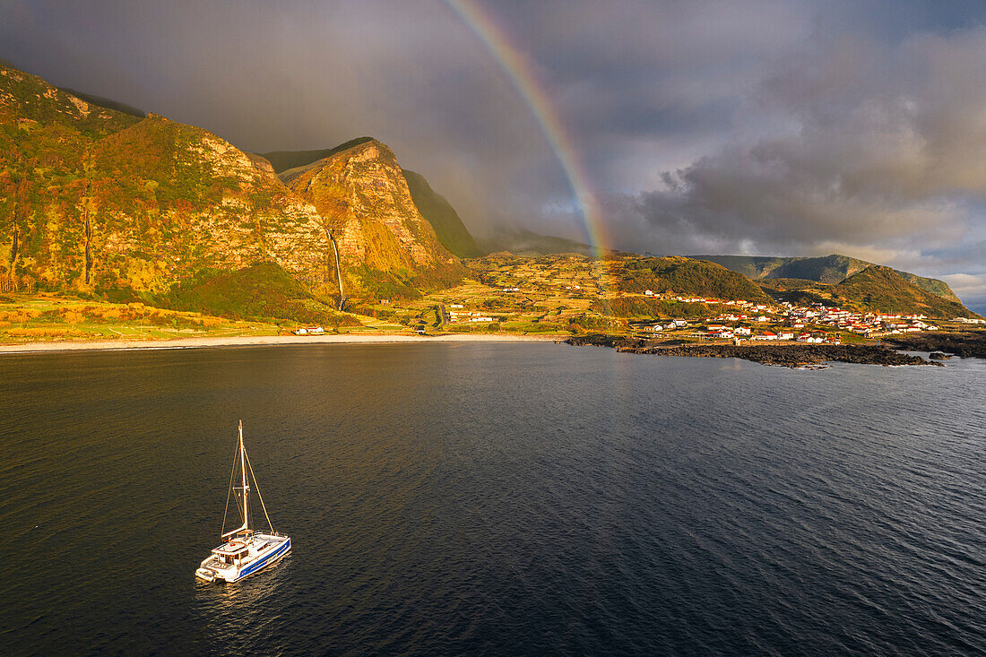 Blick auf den Sonnenuntergang am Meer von Faja Grande mit Regenbogen und Poco do Bacalhau Wasserfall im Hintergrund und Boot, Faja Grande, Lajes das Flores, Flores Insel (Ilha das Flores), Azoren Archipel, Portugal, Europa