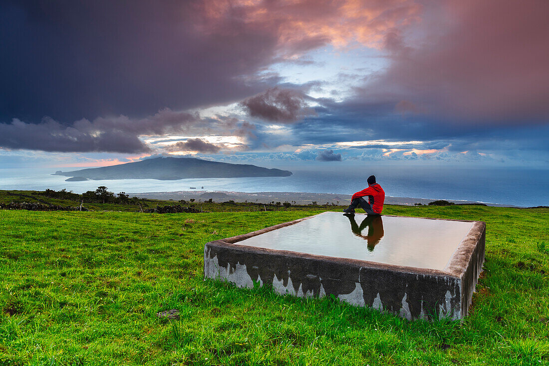 Mann sitzt auf dem Trog und bewundert die Aussicht auf die Insel Faial von der Insel Pico bei Sonnenuntergang, Gemeinde Madalena, Insel Pico (Ilha do Pico), Azoren-Archipel, Portugal, Europa