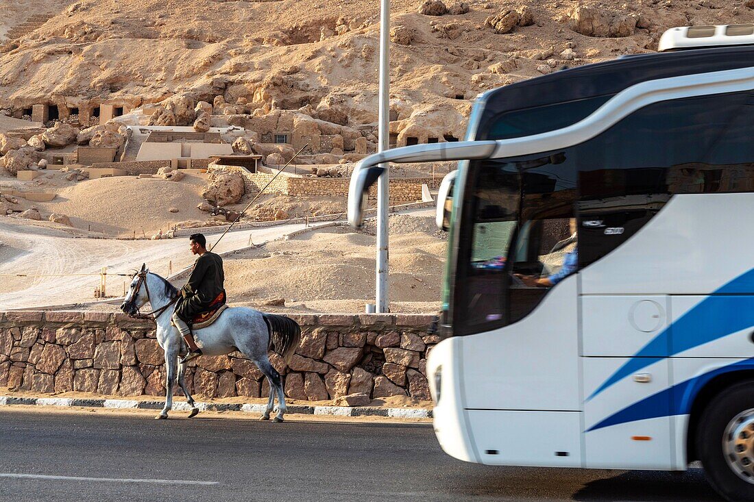 Kontrast zwischen einem Mann auf einem Pferd und einem Touristenbus vor dem Tal der Adligen, in dem sich die Gräber vieler Adliger des Neuen Reiches befinden, Luxor, Ägypten, Afrika