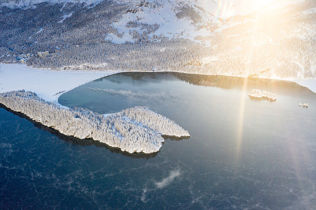 Sonnenstrahl in der Morgendämmerung auf dem zugefrorenen Silsersee und dem verschneiten Dorf Sils Maria, Kanton Graubünden, Engadin, Schweiz