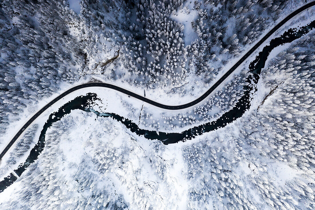 Empty snowy road running along frozen river and forest in winter, aerial view, Switzerland