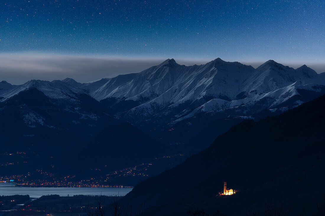 Starry night over illuminated chapel and snowcapped mountains of Alto Lario, Costiera dei Cech, Valtellina, Lombardy, Italy