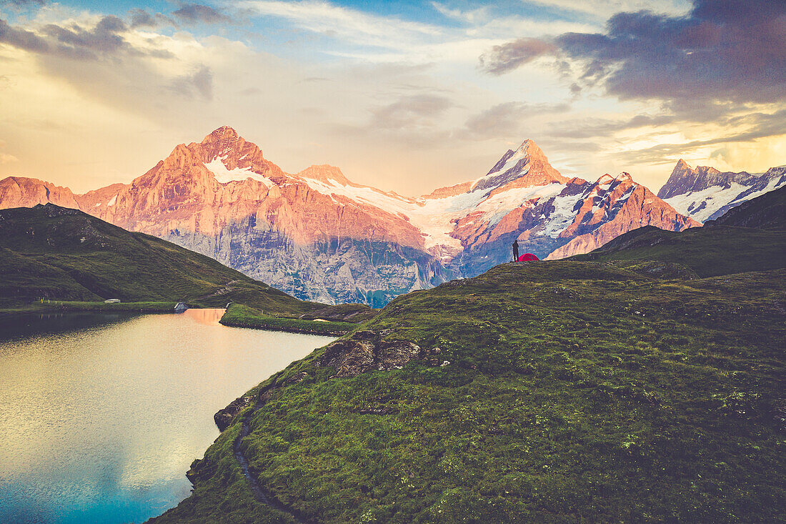 Hiker with tent looking at Bachalpsee lake and mountains at sunset, Grindelwald, Bernese Oberland, Switzerland