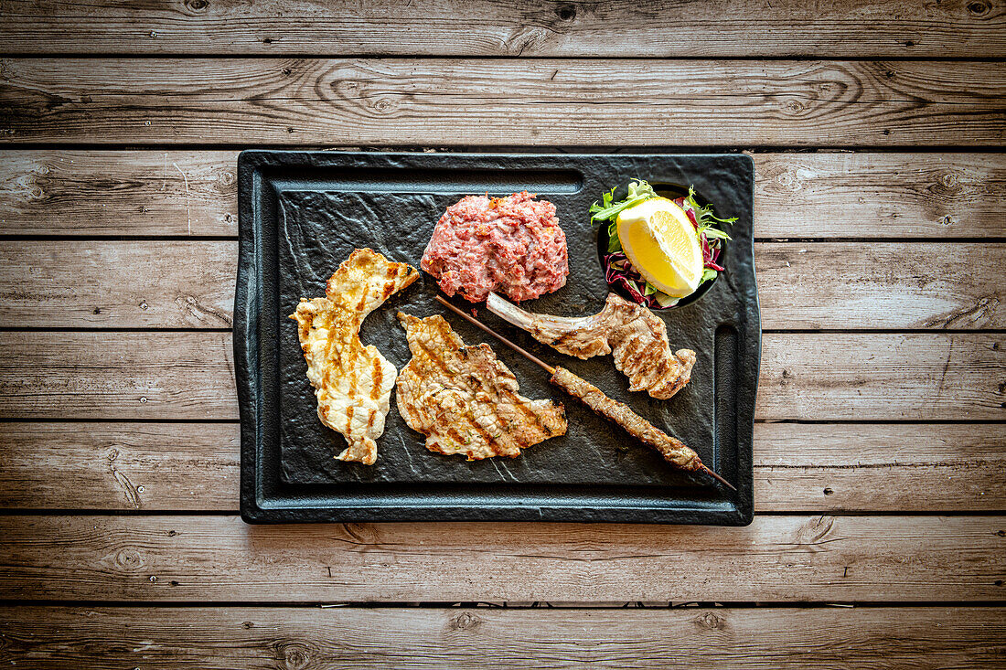 Mixed grilled meat in black tray on rustic wood table background from above, Italy