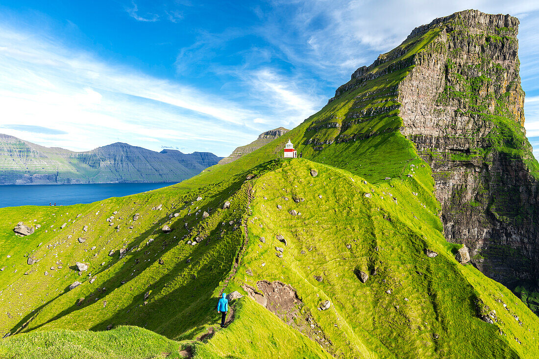 Hiker walks the green ridge of Kallur cliffs with the lighthouse in the background, Kalsoy island, Faroe islands, Denmark, Europe
