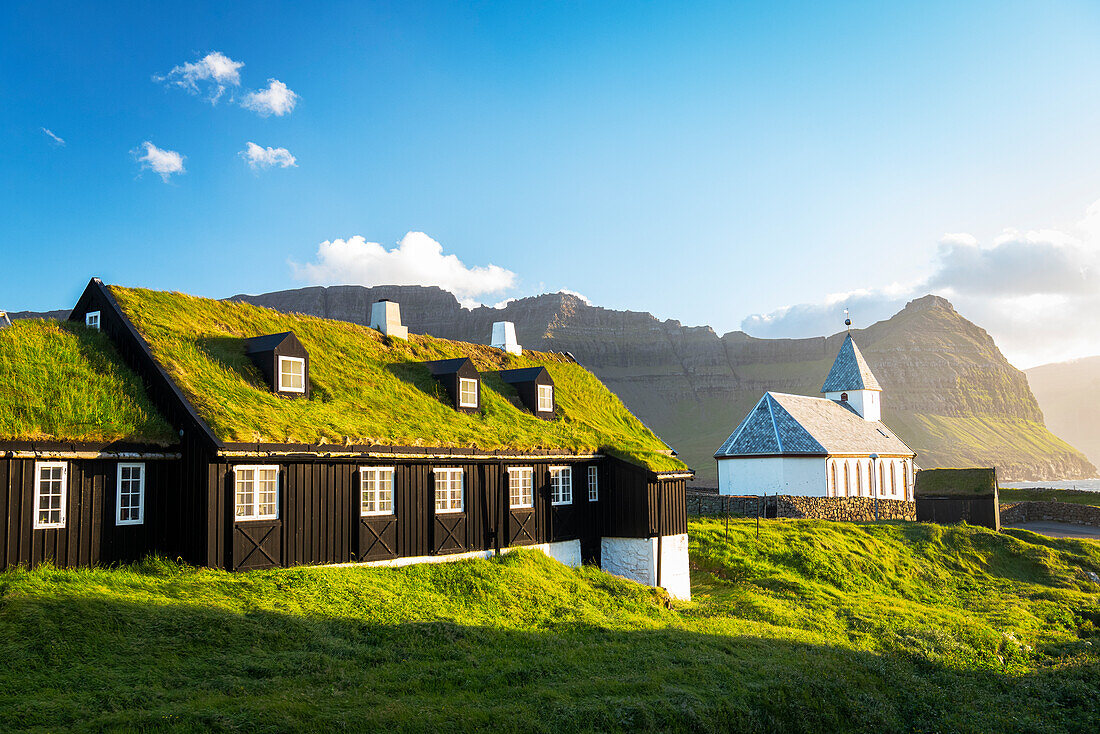 Traditionelles Gebäude mit Grasdach und die Kirche im warmen Licht des Sonnenuntergangs, Vidareidi, Insel Vidoy, Färöer Inseln, Dänemark, Europa