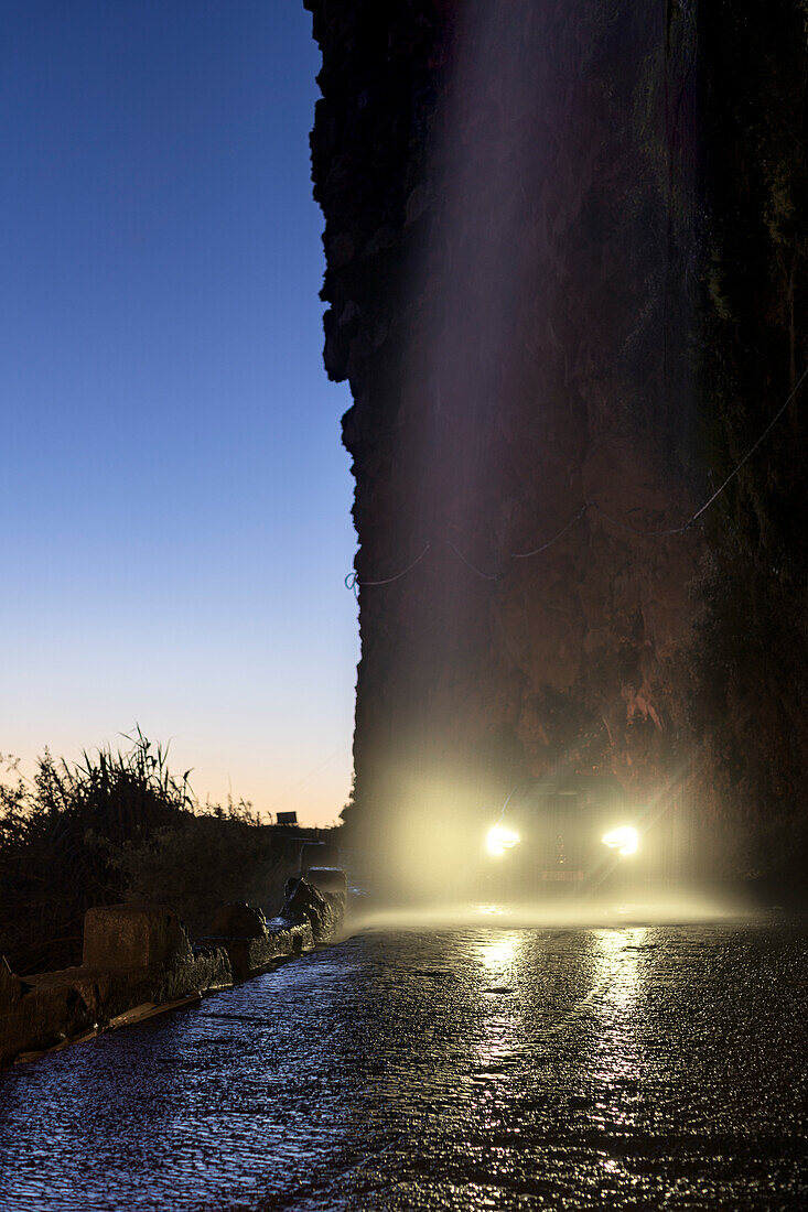 Auto fährt durch den spritzenden Anjos-Wasserfall mit eingeschalteten Scheinwerfern zur blauen Stunde, Ponta do Sol, Madeira, Portugal