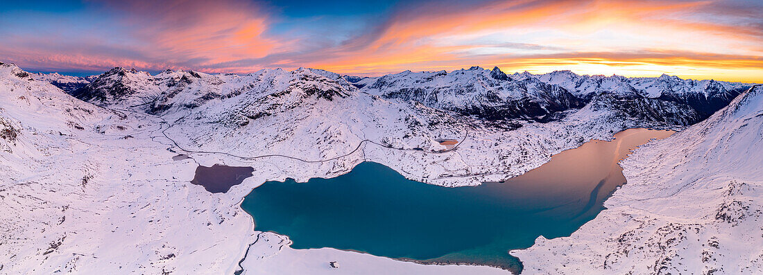 Winterlicher Sonnenaufgang über schneebedeckten Bergen, die den zugefrorenen Lago Bianco am Berninapass umgeben, Graubünden, Engadin, Schweiz