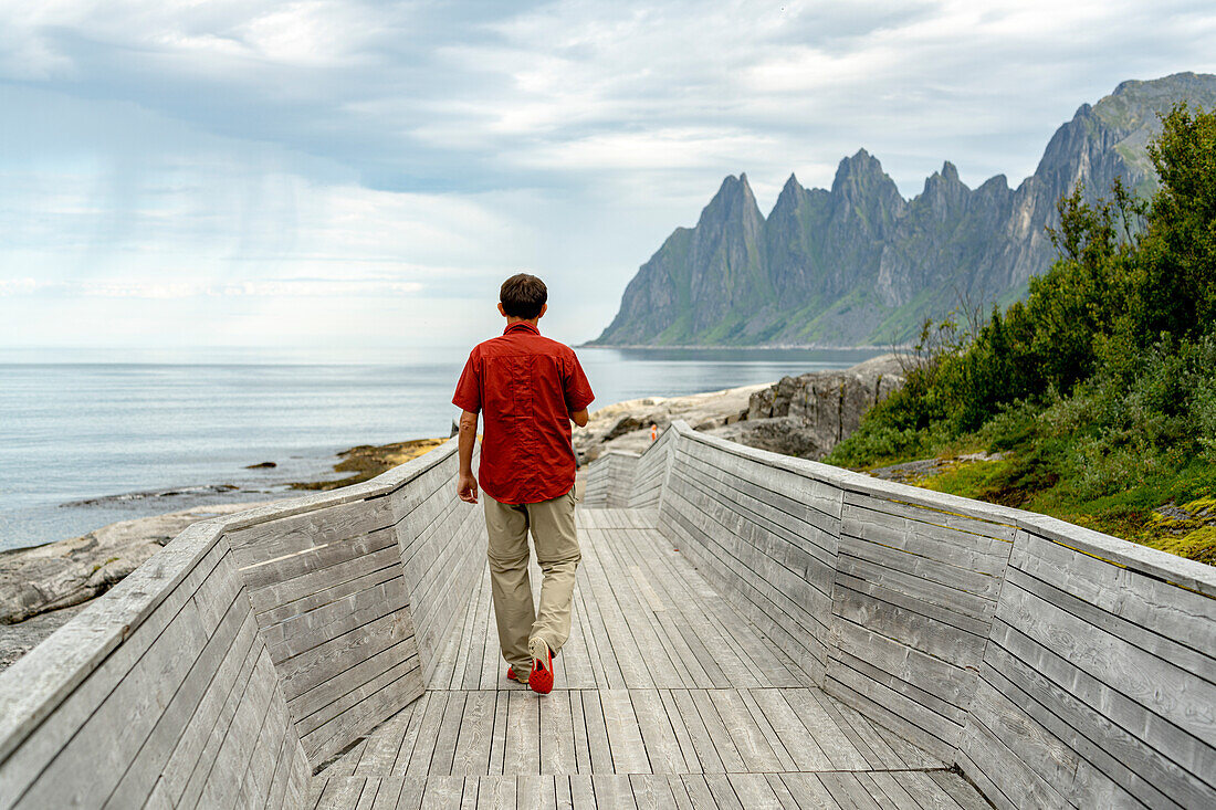 Rear view of person walking on a wooden pier to Devil's Teeth mountain peaks, Tungeneset viewpoint, Senja, Troms county, Norway