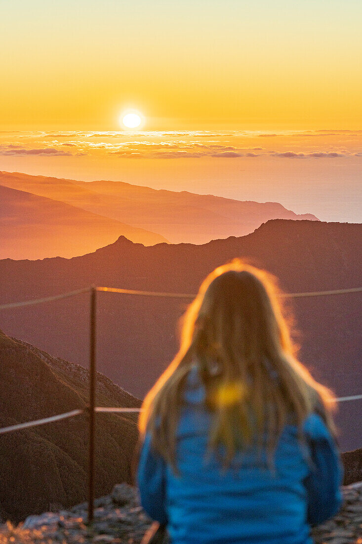 Rückansicht eines blonden Mädchens, das den Sonnenuntergang auf dem Gipfel des Pico Ruivo betrachtet, Madeira, Portugal