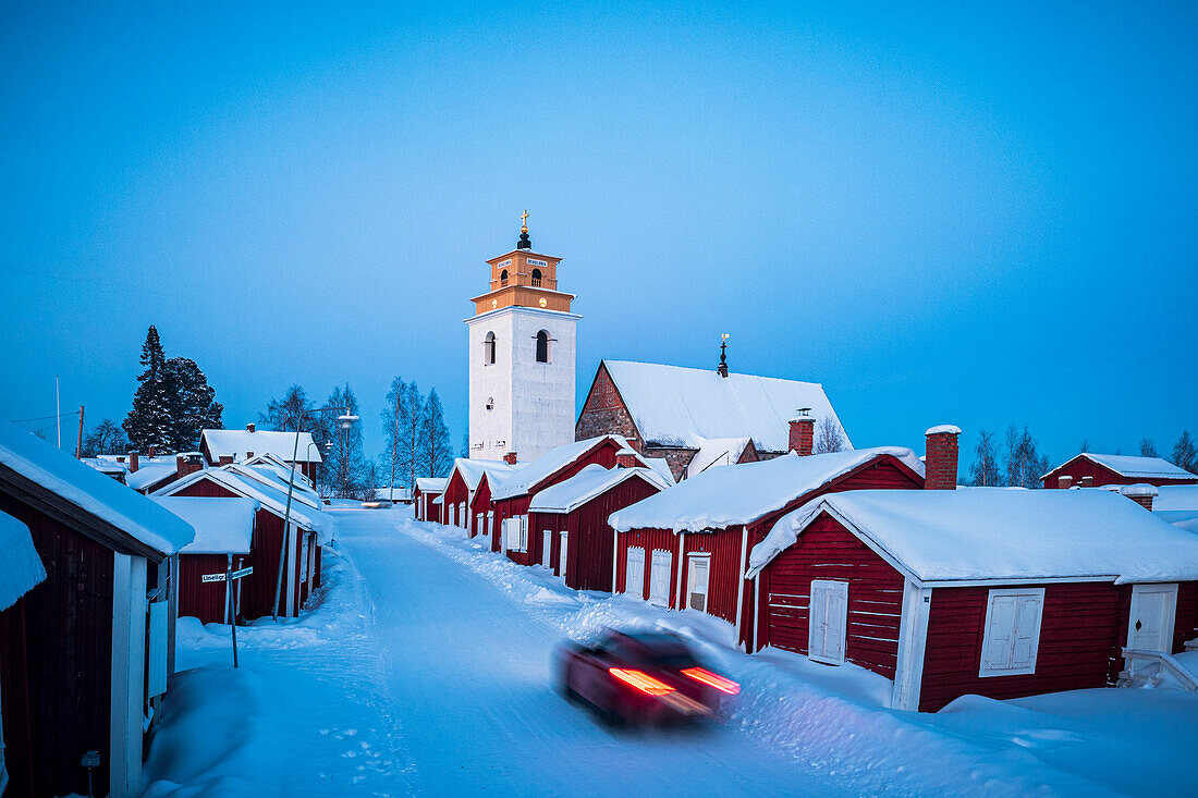 Autofahrt auf verschneiter Straße zwischen traditionellen roten Häusern in der Altstadt von Gammelstad in der Winterdämmerung, Lulea, Schweden