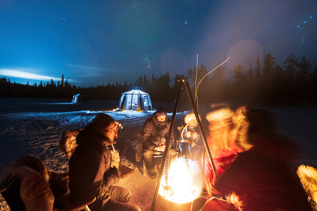 Tourists out of glass igloo warming up sitting at bonfire in the cold arctic night, Jokkmokk, Lapland, Sweden